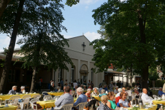 Viele Personen sitzen an den Kaffeetischen in der Sonne, im Hintergrund steht zwischen den Bäumen die Kirche.