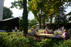 Hinter einer Hecke stehen Bänke, auf der Personen sitzen, im Hintergrund die Kaffeetische und der Pavillon zwischen Bäumen.